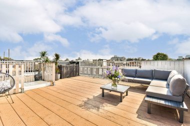 Güzel tasarlanmış, rahat oturaklı, ahşap süslemeli ve berrak mavi gökyüzüne karşı dizilmiş canlı bitkilerle dolu modern bir teras..