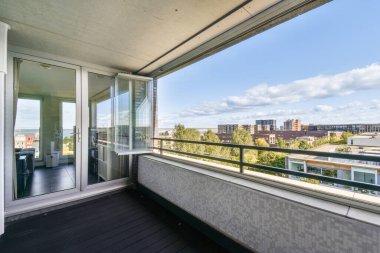 Canlı bir şehir manzarasına bakan çağdaş bir balkon, bulutlu bir gökyüzünün altında konut binaları ve yeşil alanların karışımını gösteriyor..