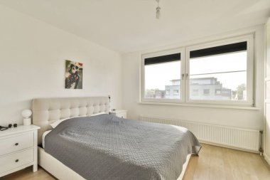 Modern bir yatak odasında minimalist iç tasarım sergileniyor. Lüks bir yatağı, parlak mobilyaları ve geniş pencereleri var. Doğal ışığın içeri girmesini sağlıyor..