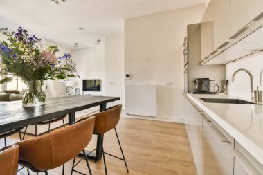 Parlak, modern bir mutfak. Parlak bir tasarım, ahşap döşeme, şık sandalyeli bir yemek masası ve dekoru yükselten canlı bir çiçek aranjmanı..