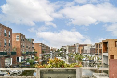 Modern konut binaları, yemyeşil yemyeşil ve bulutlu bir gökyüzünün altındaki suya renkli yansıyan sakin bir sahil manzarası..