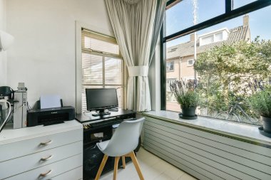 Masasında bir bilgisayar, şık bir sandalye ve bahçeye bakan geniş pencereli rahat ve işlevselliği kusursuz bir şekilde harmanlayan iyi ışıklandırılmış bir ev ofisi..