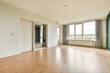 Parlak, geniş pencereli, doğal ışığın içeri girmesini sağlayan, ahşap döşemeyi ve minimalist tasarımı aydınlatan geniş bir oda..