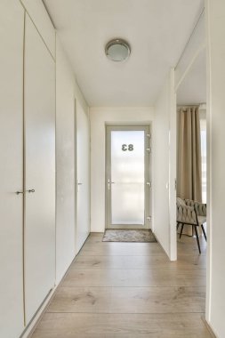 Parlak, modern koridorda parlak beyaz duvarlar, ahşap döşemeler ve şık bir kapı var. Dekor, temiz bir görünüm için minimalist tasarım unsurlarını içeriyor.