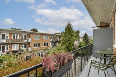 Yeşillik ve canlı çiçeklerle çerçevelenmiş modern apartmanların sergilendiği balkondan manzaralı bir manzara. Güneşli gökyüzü davetkar atmosfere ekleniyor.