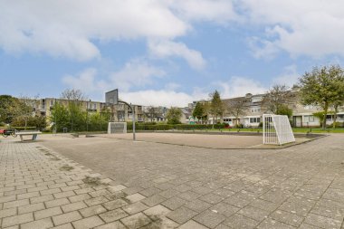 Açık mavi gökyüzü altında ağaçlar ve konut binalarıyla çevrili geniş bir şehir parkı, dinlenme ve rahatlama için ideal..