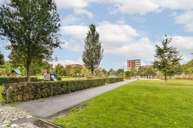 Canlı bir park, düzgünce budanmış yeşillikler, yürüyen bir patika ve etrafında bulutlu mavi gökyüzü altındaki binalarla çevrili bir çocuk oyun alanı..