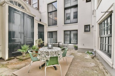 Yeşil sandalyelerle çevrili yuvarlak bir masanın yer aldığı, modern mimari öğelerin ve yumuşak yeşilliklerin arka planına karşı kurulmuş davetkar bir açık kafe alanı..