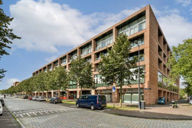 Çağdaş bir binada, verimli yeşillik ve ağaçlarla çevrili geniş cam pencereler park halindeki araçlarla kalabalık bir şehir caddesinde yer alıyor..