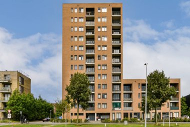 Bulutlu bir gökyüzü ve yeşil bir çevreye karşı kurulmuş, birden fazla balkonlu kahverengi tuğlalardan yapılmış uzun, modern bir yerleşim yeri..