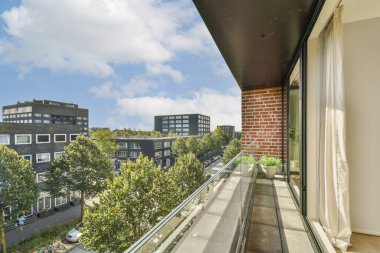 Parlak bir gökyüzü altında yeşillik ve çağdaş mimariyle süslenmiş hareketli bir şehir caddesine bakan modern bir balkondan davetkar bir manzara..