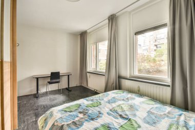 Geniş pencereli, basit dekorlu, parlak, minimalist bir oda ve uzayın huzurlu atmosferini arttıran renkli bir yatak örtüsü..