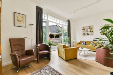 Şık mobilyaların ve geniş pencerelerin bulunduğu modern bir oturma odası yeterli miktarda doğal ışığı içeri alarak sıcak ve davetkar bir atmosfer yaratıyor..