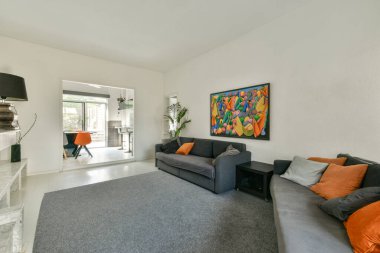 Gri bir kanepe, renkli bir duvar sanatı ve parlak turuncu bir sandalye içeren şık bir oturma odası, canlı ve çağdaş bir atmosfer yaratıyor..