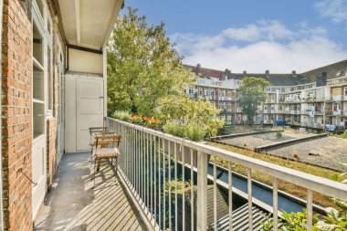 Güneşli bir günde huzurlu bir şehir avlusundaki büyüleyici binalar tarafından çerçevelenmiş, yemyeşil ve rahat koltuklar sergilenen sakin bir balkon manzarası..