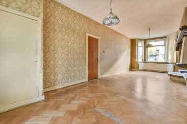 Dekoratif lambaları, büyük pencereleri ve desenli duvar kağıtlarıyla kaplı duvarları olan boş bir oda.