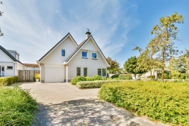 Arka planda beyaz kenarlı ve mavi gökyüzü olan bir ev var. Ön kapıya giden bir garaj yolu var.