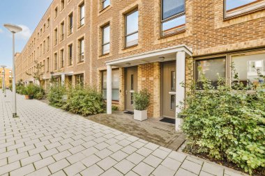 Her iki tarafında bitkiler ve çalılar olan tuğla bir bina. Kapının yanında sokak lambası direği var.