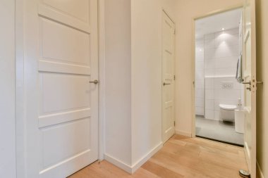 Beyaz kapıları olan, çağdaş bir banyoya çıkan, gösterişli, modern bir koridor. Minimalist tasarım, temiz çizgiler ve nötr tonları vurgular..