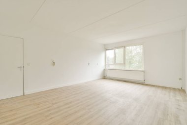 Geniş ve parlak bir oda, parlak ahşap döşeme, beyaz duvarlar ve geniş bir pencere, doğal ışığın boşluğu doldurmasına izin veriyor..