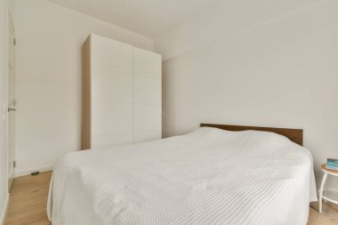 Temiz ve modern bir yatak odası, minimalist tasarım unsurları sergiliyor, beyaz bir battaniye ile kaplı basit bir yatak ve dağınık görünümü için parlak mobilyalar..