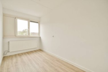 Parlak ve minimalist bir iç mekan düz beyaz bir duvar, ahşap döşeme ve doğal ışığın içeri girmesine izin veren geniş bir pencere, havadar bir atmosfer yaratıyor..