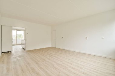 Geniş, boş bir oda. Açık renkli ahşap döşeme ve beyaz duvarlar var. Camlardan içeri ışık süzülen sakin ve minimalist bir estetik yaratıyor..