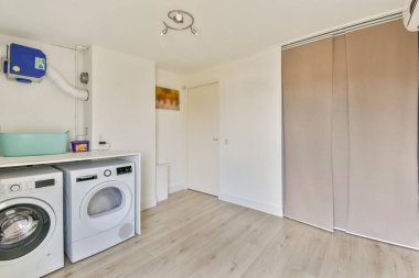 Açık ahşap döşeme ve nötr renklerle bir çamaşır makinesi, kurutma makinesi ve organize bir alan içeren temiz ve minimalist bir çamaşır odası..