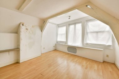 Büyük pencereleri, soluk ahşap döşemeleri ve ince duvar kusurları olan, eşsiz bir atmosfer yaratan boş, güneşli bir minimalist oda..