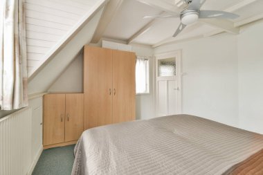 Görüntüde şık bir yatak odası, düzgün yapılmış bir yatak odası, ahşap gardırop ve tavan vantilatörü var. Huzurlu ve davetkar bir ortam yaratıyor..