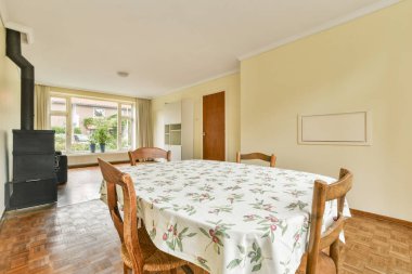 Çiçekli bir masa örtüsü, ahşap döşeme ve bahçe manzarası sunan geniş pencereli parlak bir yemek alanı sıcak ve davetkar bir atmosfer yaratıyor..