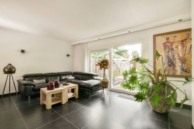 Rahat bir oturma odası, dekoratif bitkiler ve doğal ışık getiren büyük pencereler, sıcak bir ortam yaratıyor..