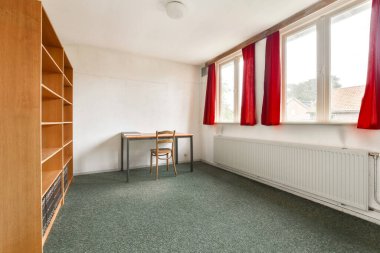 Tahta bir masa, bir sandalye ve tarafsız duvarlar ve halıyla çelişen parlak kırmızı perdelerin yer aldığı basit ve temiz bir iç mekan..