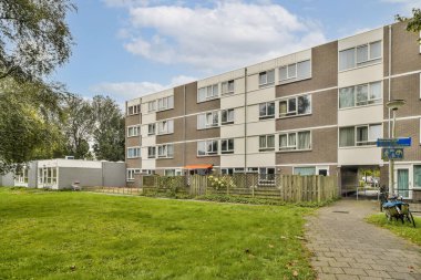 Çağdaş bir konut binası. Birden fazla penceresi var. Çimenli bir alan ve patikaların yanında yer alıyor..