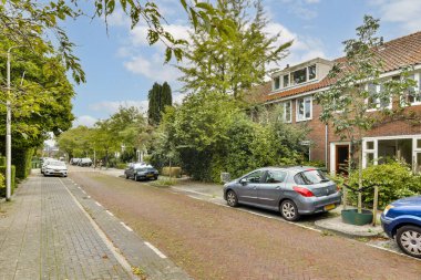 Huzurlu bir banliyö caddesi ağaçlar ve evlerle dolu, huzurlu bir yerleşim alanı sergiliyor. Arabalar kaldırım taşı yolu boyunca açık gökyüzünün altında park halinde..