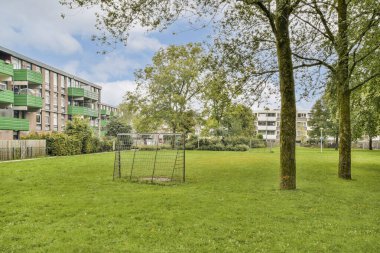 Çağdaş apartman binaları tarafından çerçevelenmiş geniş yeşil çimenler ve olgun ağaçlar sergilenen canlı bir şehir parkı..