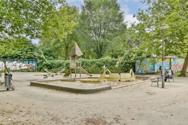 Sahnede oyun yapısına, banklara ve uzun ağaçların ve parlak gökyüzünün ortasındaki renkli duvar resimlerine sahip bir oyun alanı ve canlı bir atmosfer yaratıyor..