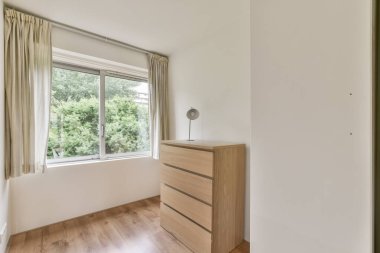 Parlak ve havadar bir odada ahşap bir şifonyer ve yeşilliğe tepeden bakan geniş bir pencere, minimalist bir ev tasarımı somutlaştırılmış..