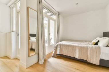 Geniş bir ayna, yumuşak yatak örtüsü ve geniş pencerelerden gelen doğal ışık içeren şık ve parlak bir yatak odası, sakin bir atmosfer yaratıyor..