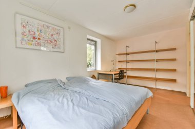 Parlak ve ferah bir yatak odası, düzgün yapılmış bir yatak odası, bir masa ve basit bir raf. Dekor minimalist, yumuşak doğal ışık sükuneti arttırıyor..