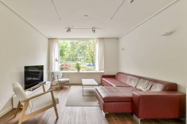 Kırmızı deri bir kanepe, minimalist mobilyalar ve geniş pencereli şık modern bir oturma odası. Doğal ışığın içeri girmesini sağlıyor. Hoş bir atmosfer yaratıyor..