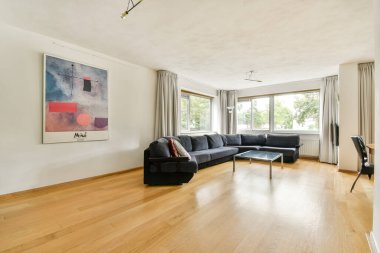 Büyük bir kanepe, ahşap döşeme ve pencerelerden gelen parlak doğal ışık altında duvarda renkli bir sanat eserinin yer aldığı şık ve ferah bir oturma odası..