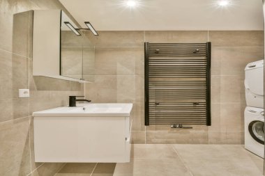 Çağdaş eşyalar, geniş bir lavabo, zarif fayanslar ve modern ama işlevsel bir yer yaratan şık bir havlu radyatörü olan parlak bir banyo..