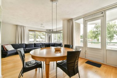 Geniş, modern bir oturma odasında yuvarlak bir yemek masası, rahat koltuklar ve doğal ışık ve yeşillik manzarası sunan geniş pencereler bulunur..