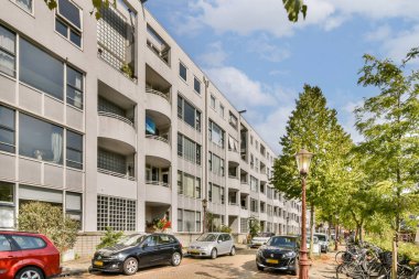 Çağdaş mimari tasarımı yansıtan, yeşillik ve park halindeki arabalarla çevrili, modern bir şehir apartmanını gösteriyor..