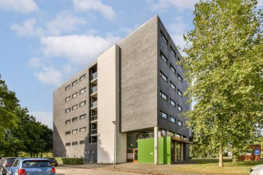 Çağdaş bir bina, parlak yeşil ağaçlardan oluşan bir zemin ve modern mimariyi gözler önüne seren parlak bir gökyüzü üzerine kurulu..