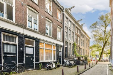 Amsterdam 'ın pitoresk caddesinde klasik tuğla binalar, bisikletler ve şehrin zengin mimari mirasını temsil eden güneşli bir gökyüzü sergileniyor..