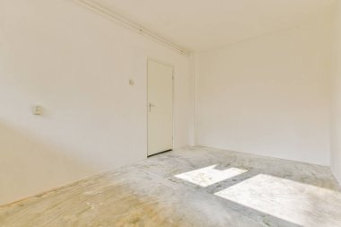 Çıplak duvarlar, bir kapı ve güneş ışığının yer yüzüne gölge düşürdüğü parlak ve havadar boş bir oda, sakin, minimalist bir alan yaratıyor..