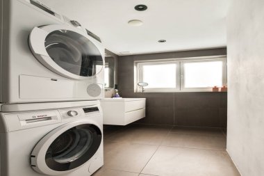 Çağdaş fayanslar ve parlak doğal ışık ile tamamlanmış, minimalist tasarımı ile üst üste dizilmiş çamaşır ve kurutma makinelerinin yer aldığı şık ve modern bir çamaşır odası..