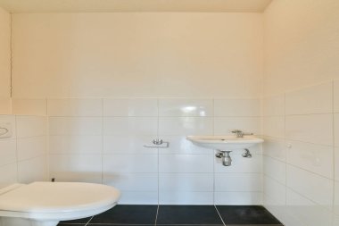 Beyaz bir duvar, siyah döşemeler ve temel konforlar, basitlik ve işlevselliği vurgulayan modern bir banyonun temiz ve minimal bir görüntüsü..